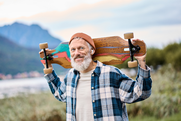 Older happy hipster man skater standing in nature park holding skate.