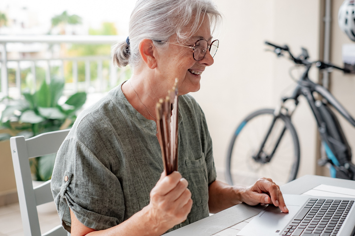 Video call concept. Smiling elderly woman having video call on laptop sitting outdoors on terrace while she fanning herself with a fan, elderly woman using modern technology and wireless connection