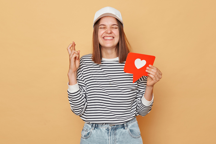 Hopeful woman wearing striped shirt and baseball cap holding like blogger icon standing isolated over beige background crossing fingers dreaming to get a lot of likes on social networks