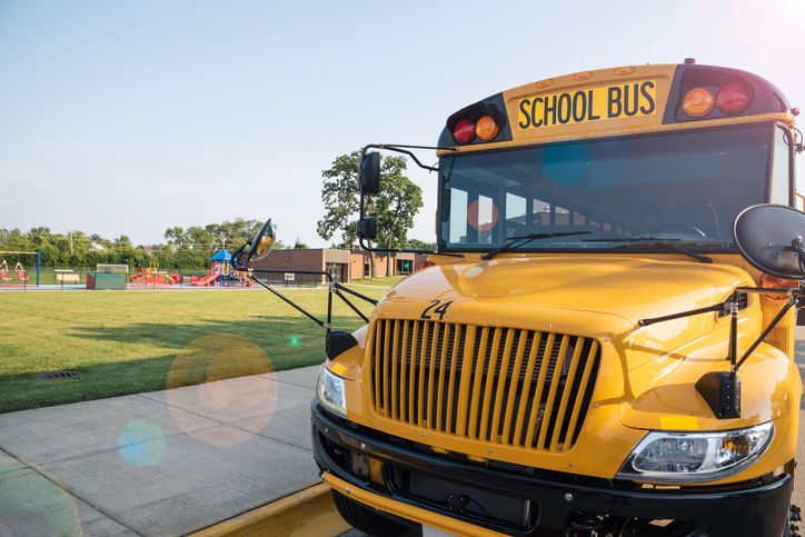 Front View Of Yellow School Bus Parked Along Sidewalk In Front Of School Playground