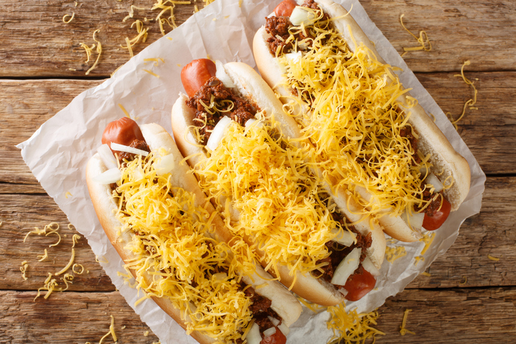 Beef hot dog in a bun covered with Cincinnati Chili, diced onions, and a mound of shredded cheddar cheese closeup in the paper. Horizontal top view
