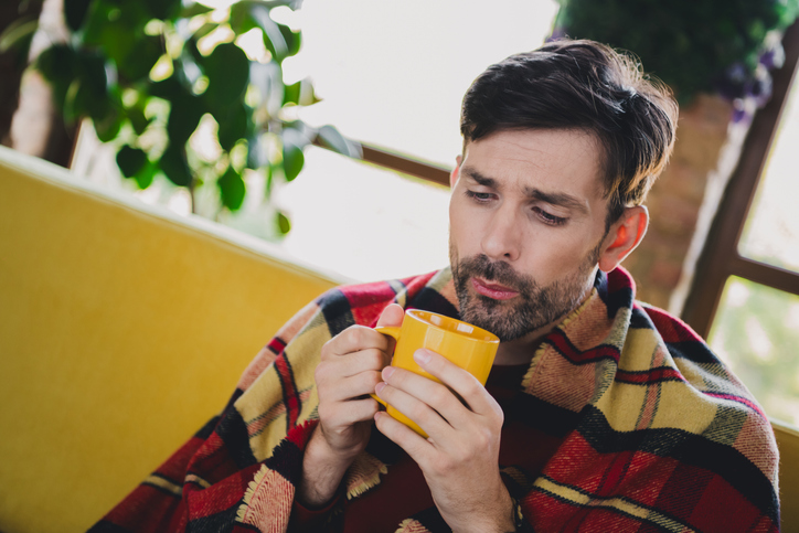 Young handsome man enjoying a relaxing weekend at home, covered with a blanket and holding a warm drink in his comfortable apartment