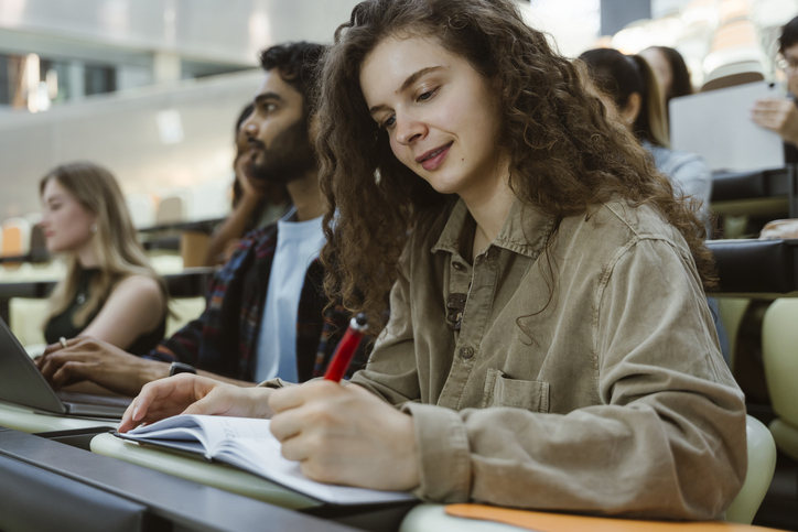 Curly haired young woman writing in book while sitting in classroom at college
