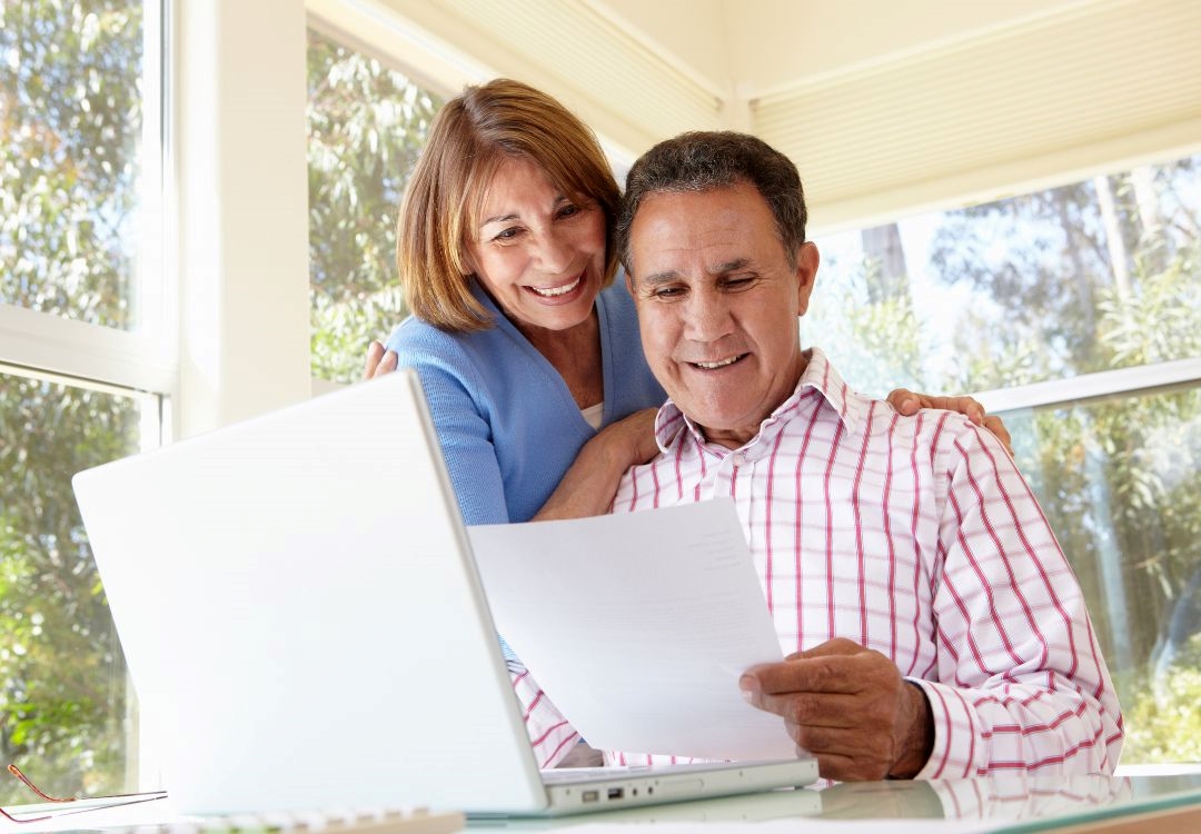 A smiling middle-aged couple sitting at a laptop computer together in a bright, sunny room.