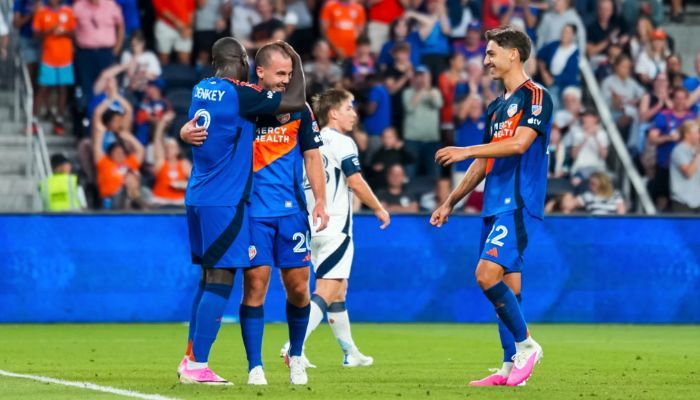 Two soccer players in blue jerseys celebrating on a soccer field with other players in the background.