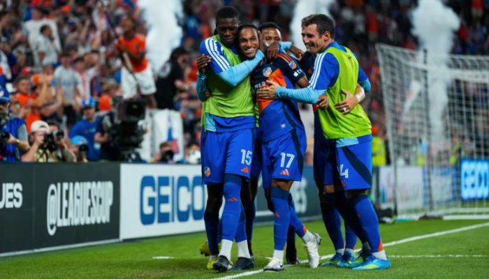 A group of soccer players celebrating a goal, with players in blue jerseys embracing and surrounded by spectators.