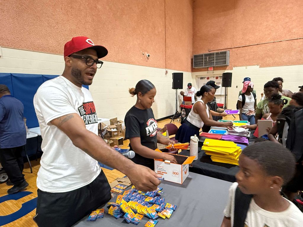 A man in a red baseball cap and white shirt is distributing school supplies to a group of children in a gymnasium.