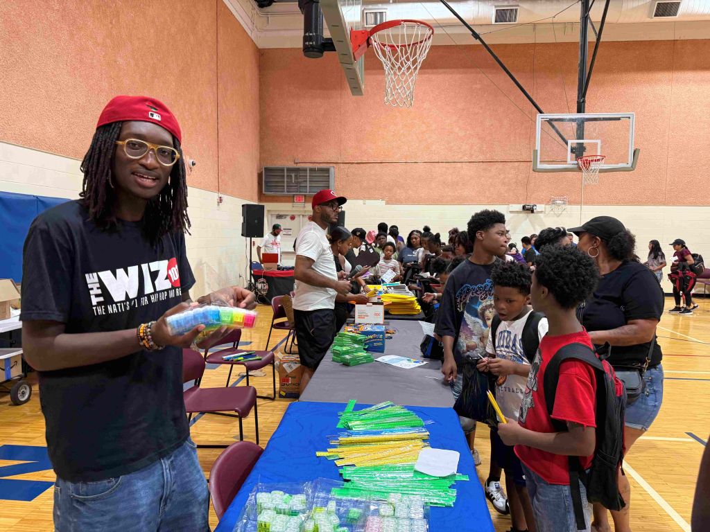 A group of people, mostly children, gathered in a basketball court. A man wearing a red cap and a t-shirt that says "The Wiz" is standing at a table distributing supplies. Basketball hoops and backboards are visible in the background.