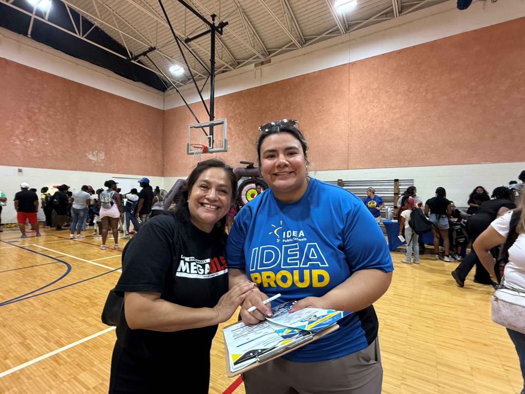 Two smiling women, one wearing a "Mega" shirt and the other wearing an "IDEA Proud" shirt, standing in a basketball gym.