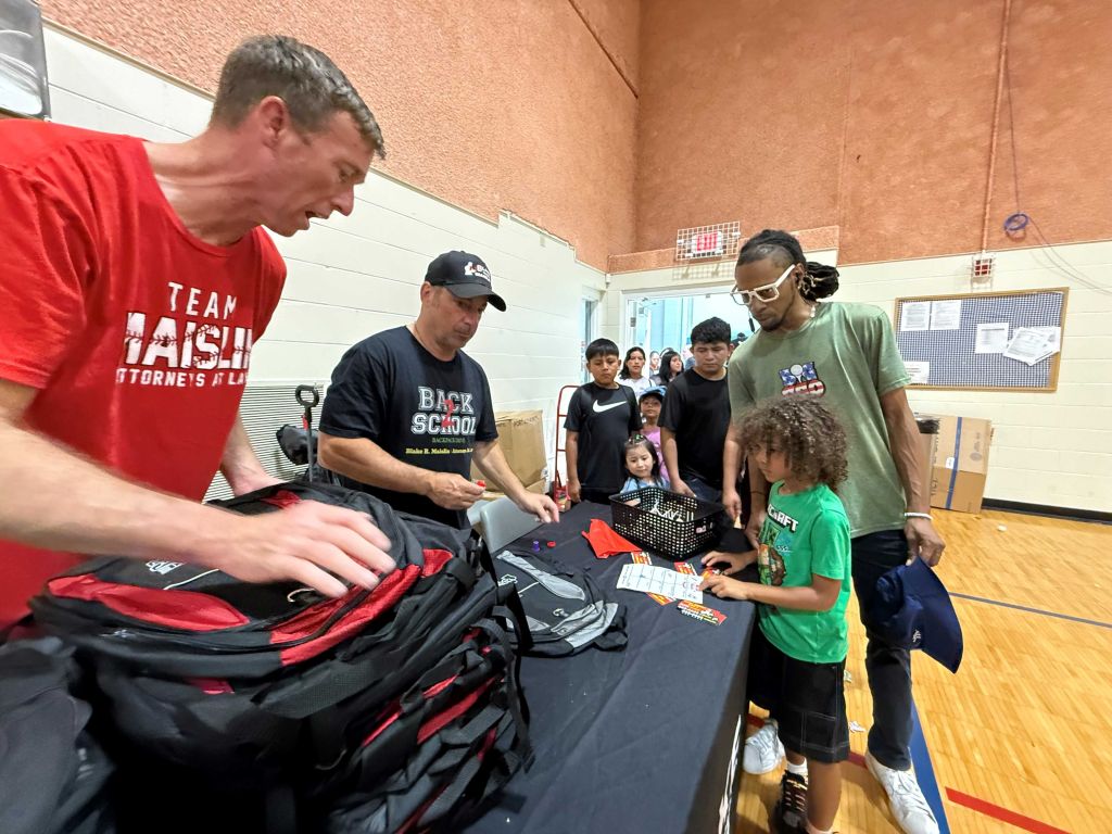 A group of people, including adults and children, gathered around a table with sports equipment and supplies. The adults appear to be distributing or organizing the items.
