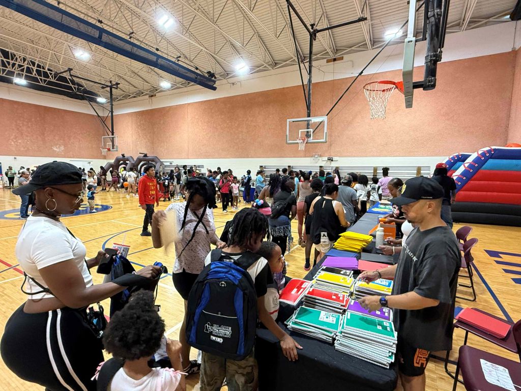 A crowded indoor basketball court with people gathered around tables displaying school supplies and other items.