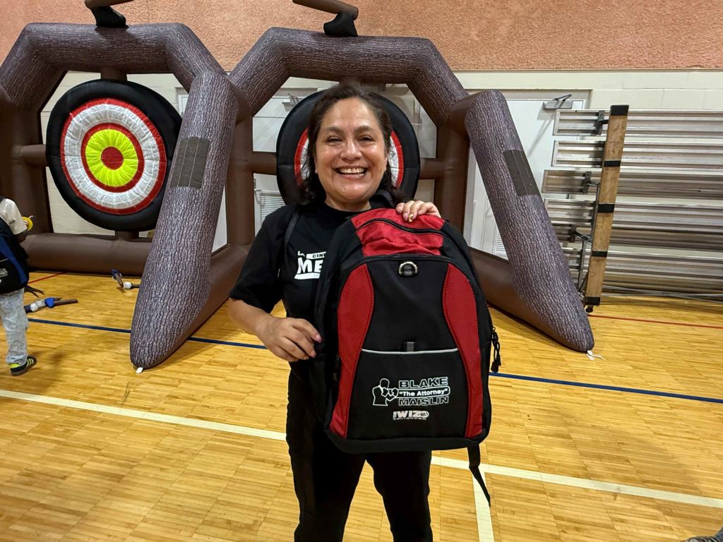A smiling woman wearing a backpack with "Blake Martin" printed on it, standing in front of archery targets in a gym.