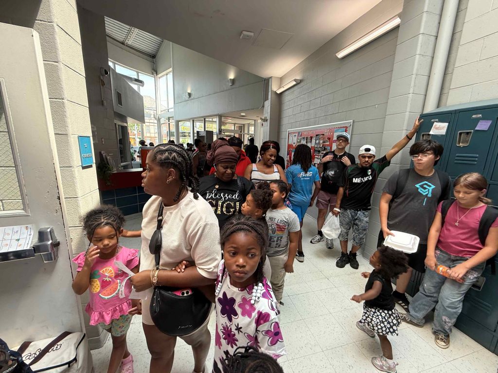 A crowded school hallway with students of various ages and backgrounds, some wearing branded clothing and backpacks.