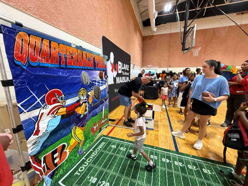 A basketball court with a large mural depicting a football player. Several people are gathered around the court, some interacting with a display or activity.