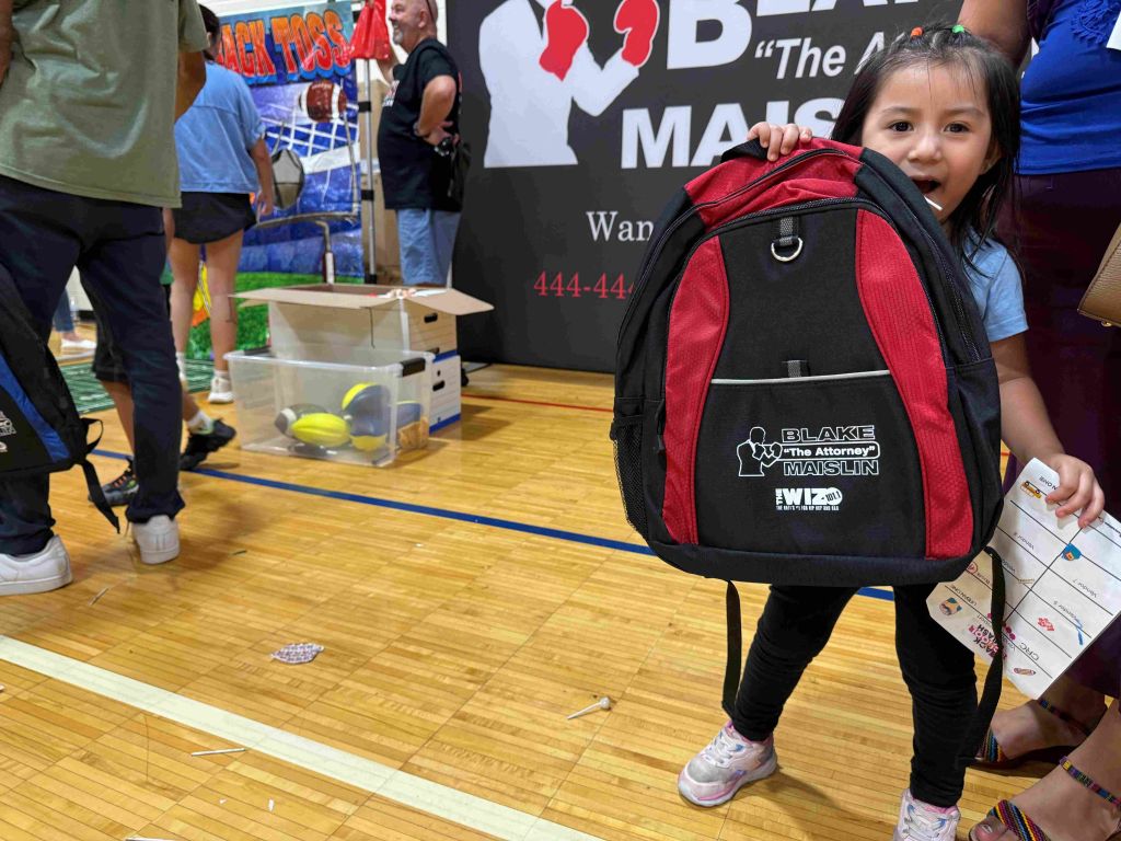 A young girl wearing a red and black backpack with the "Blake Maislin" logo stands in a room with various items and signage.