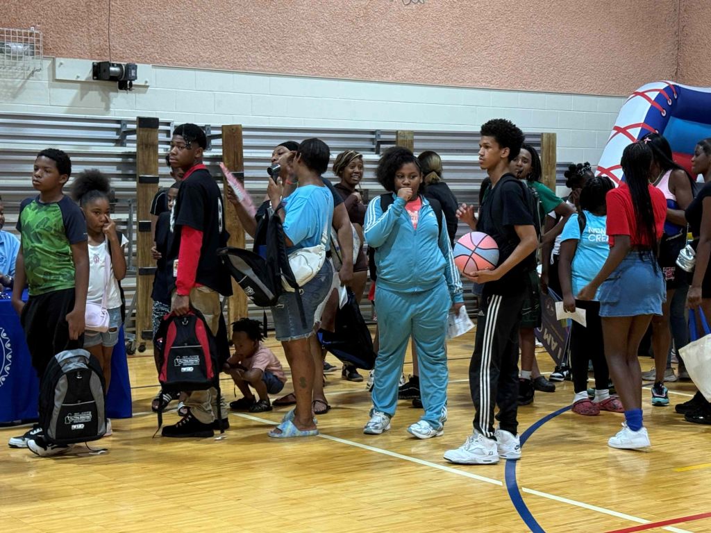 A group of young people, mostly children, gathered in a gymnasium or sports facility, some holding basketballs.