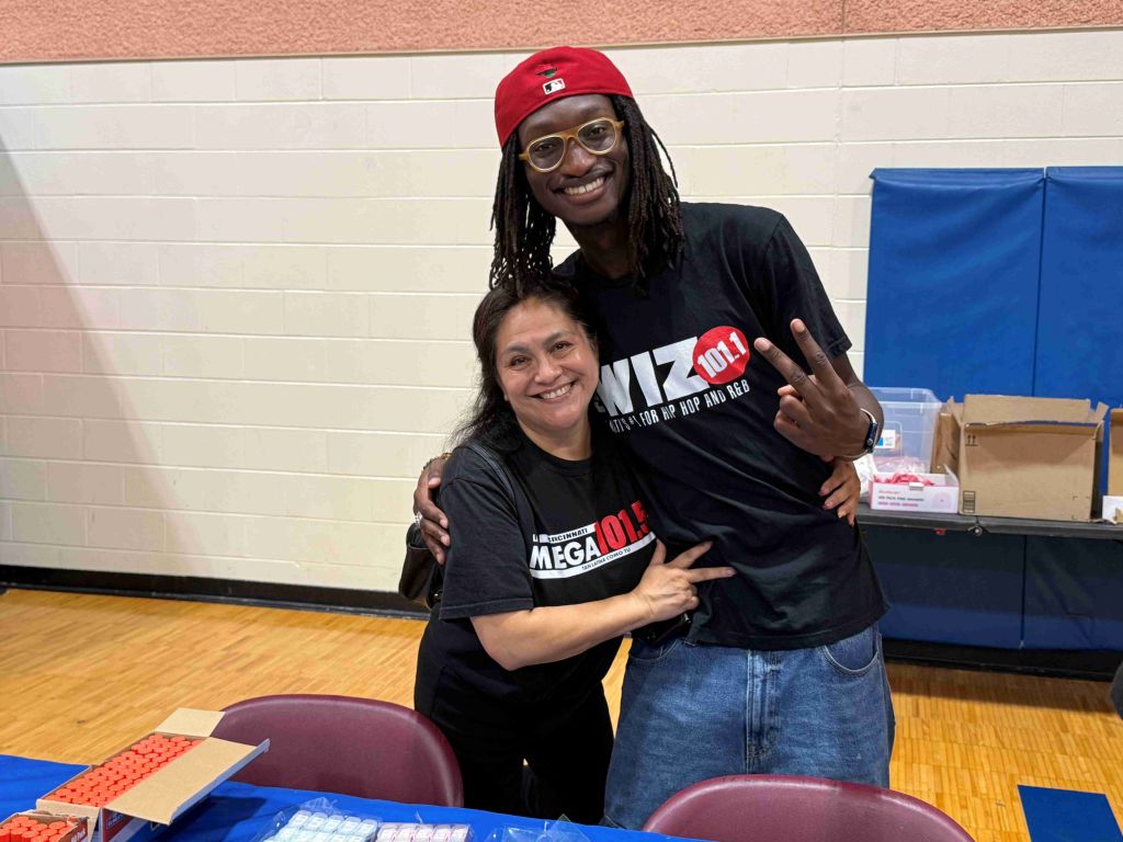 Two people smiling and embracing, one wearing a red baseball cap and the other a black t-shirt with "MEGA 101" printed on it.