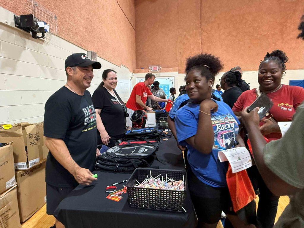 A group of people, including adults and children, gathered around a table with various items and supplies. The image shows a community event or activity.