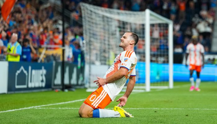 A soccer player in an orange jersey kneeling on the field, celebrating after scoring a goal.