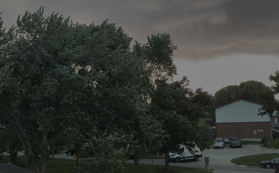 A stormy, overcast sky with lush green trees and a building in the background. Several vehicles are parked in the lot.