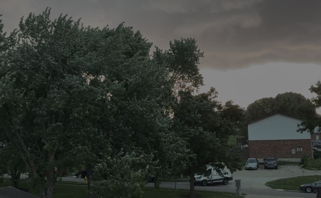 A stormy, overcast sky with lush green trees and a building in the background. Several vehicles are parked in the lot.