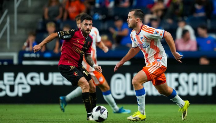 Two soccer players competing for the ball on a soccer field, one wearing a red jersey and the other wearing an orange jersey.