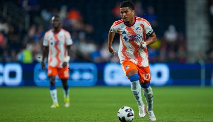 A soccer player in an orange jersey dribbling the ball on a soccer field at night.