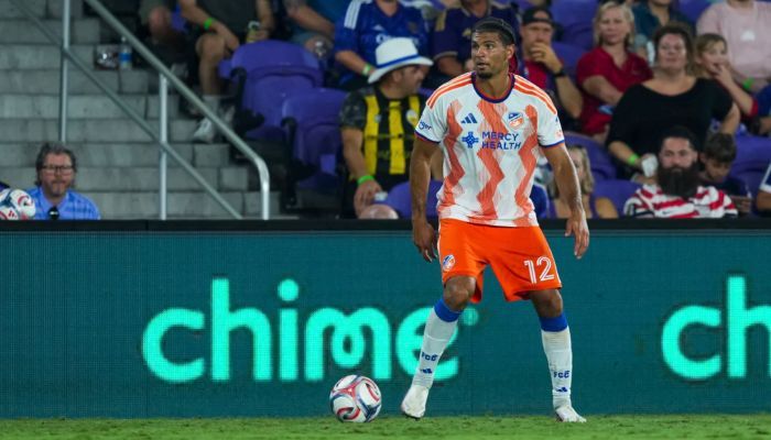A soccer player in an orange jersey dribbling the ball on a soccer field, with spectators in the background.