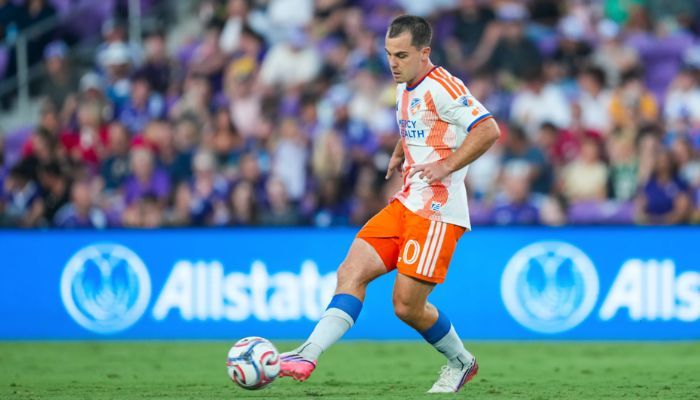 A soccer player in an orange jersey dribbling a ball on a soccer field with spectators in the background.