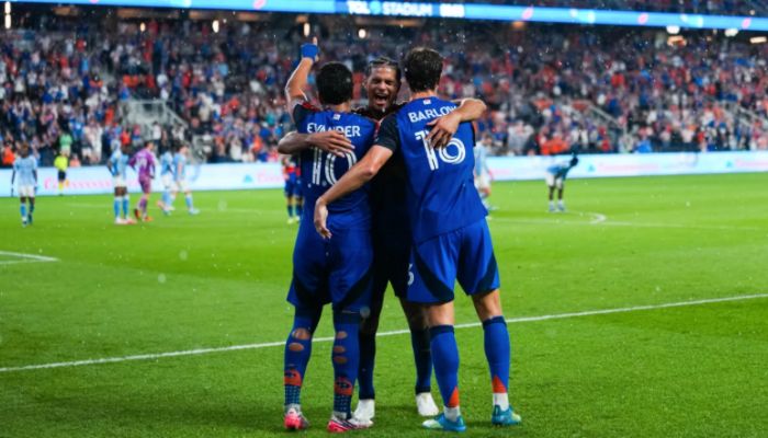 Three soccer players in blue uniforms celebrating a goal on a soccer field.