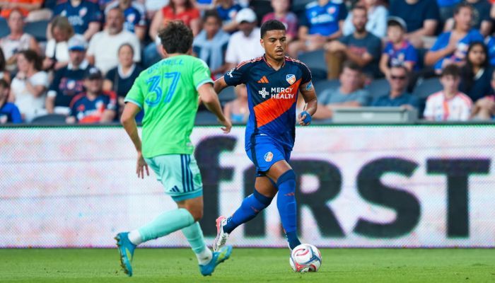 Two soccer players in action on a field, one wearing a green jersey and the other a blue jersey with "Mercy Health" printed on it.