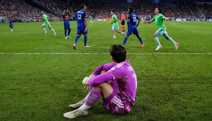 A soccer player in a purple jersey sits on the field, dejected, as the opposing team continues play on the field behind them.