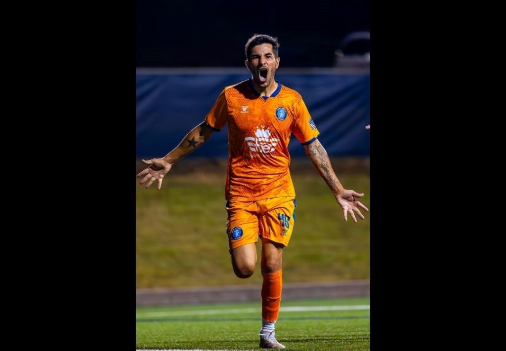 A soccer player in an orange jersey with the text "Lan Susho" on the front, celebrating with arms outstretched on a soccer field at night.