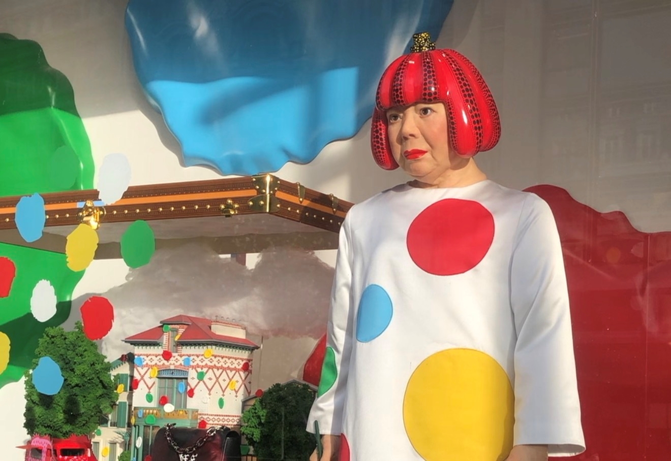 A person wearing a colorful costume with polka dots and a large red hat stands in front of a backdrop with colorful shapes and patterns.