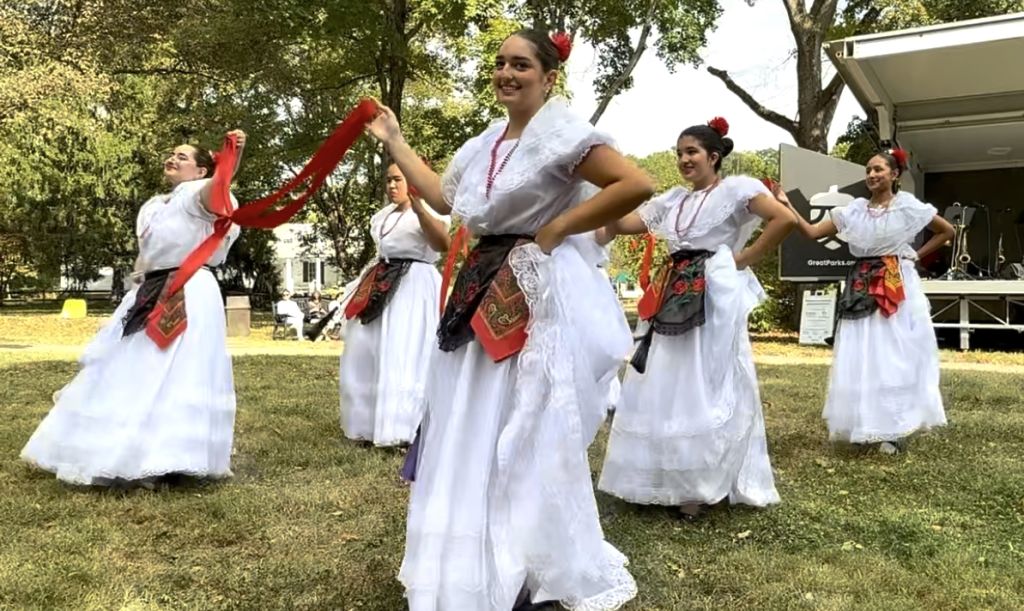 A group of women in traditional Spanish-style dresses performing a lively dance routine outdoors on a grassy field.